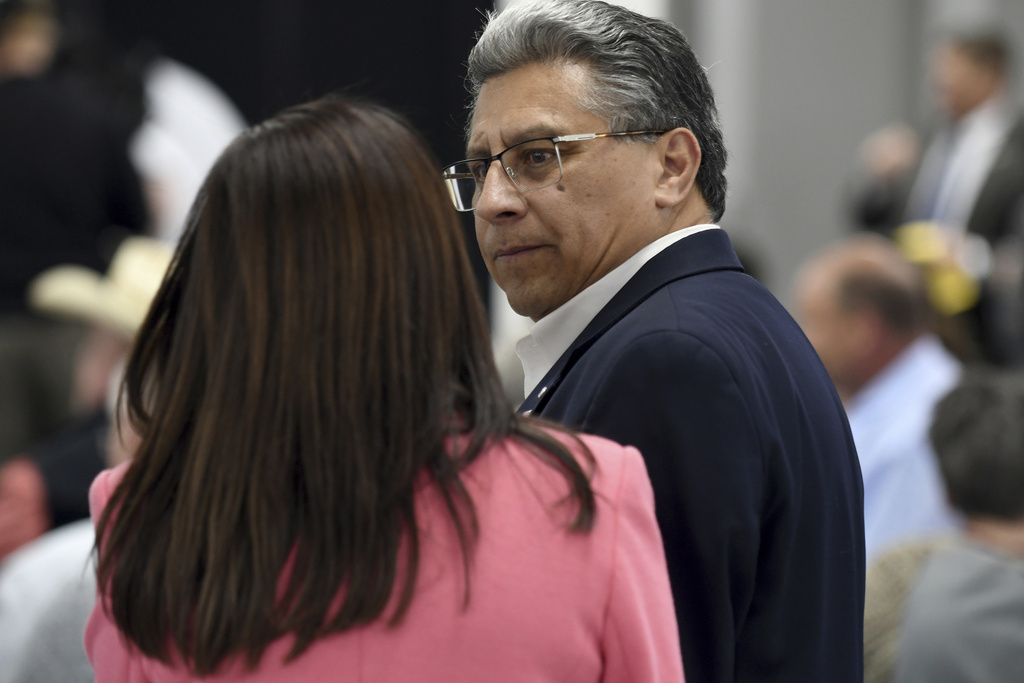 A man and a woman have a conversation in a crowded room. The man is wearing glasses and a suit, while the woman is wearing a pink jacket.