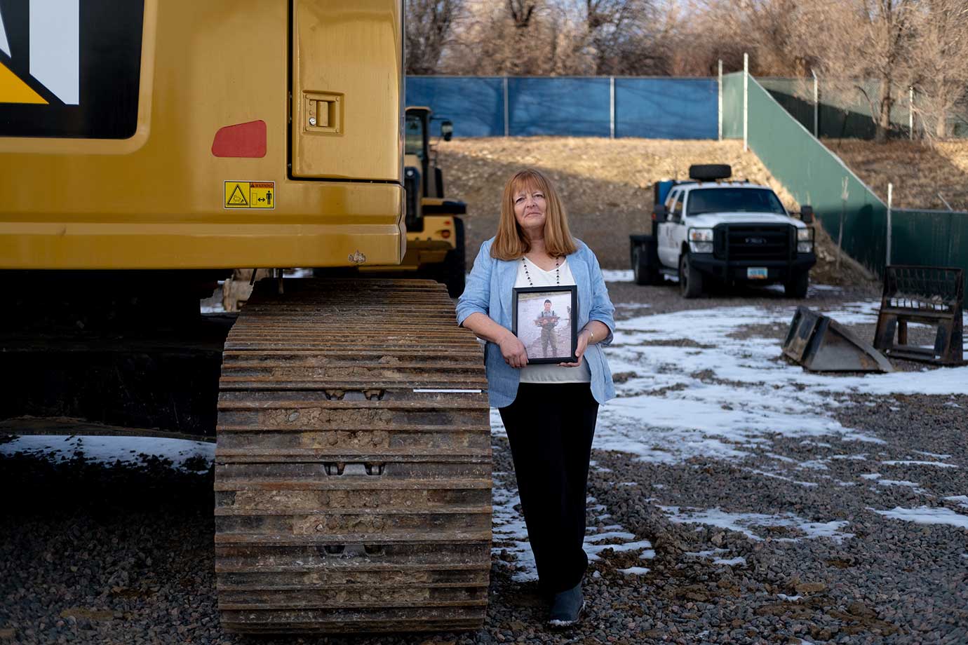 A woman stands next to a yellow excavator holding a photo of her son