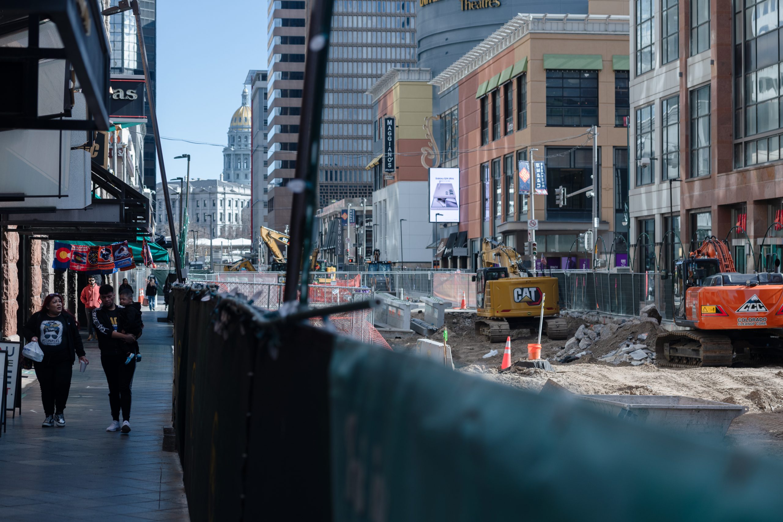City street under construction with heavy machinery and barriers. People walking on the sidewalk to the left, buildings and a distant dome visible in the background.