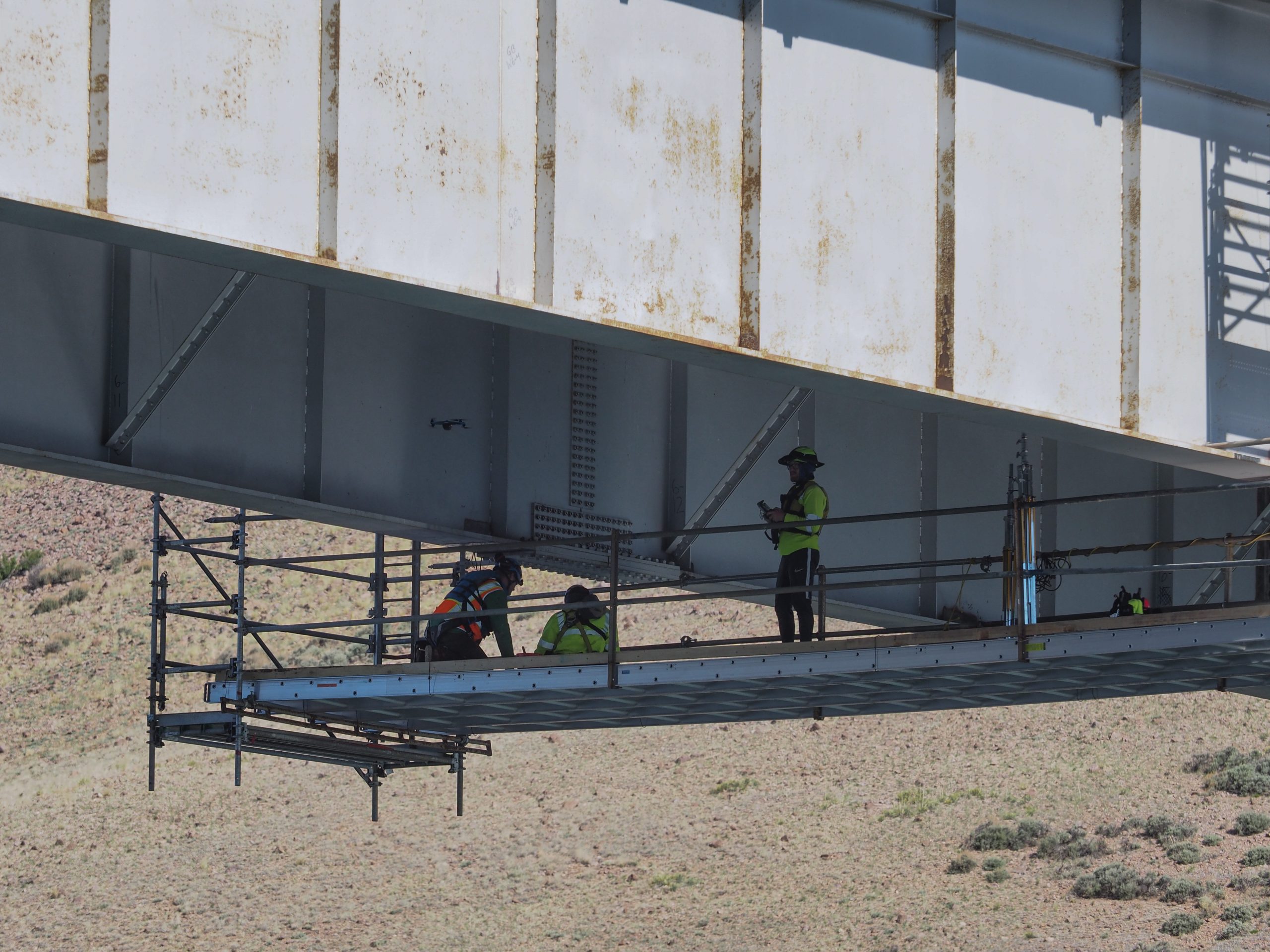 Three people in bright vests and helmets stand on scaffolding that runs under a bridge.