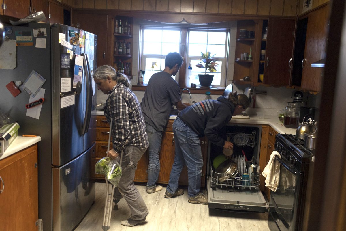 Three people are cleaning a kitchen: one is unloading a dishwasher, another is at the sink, and the third is using crutches and holding groceries near the refrigerator.