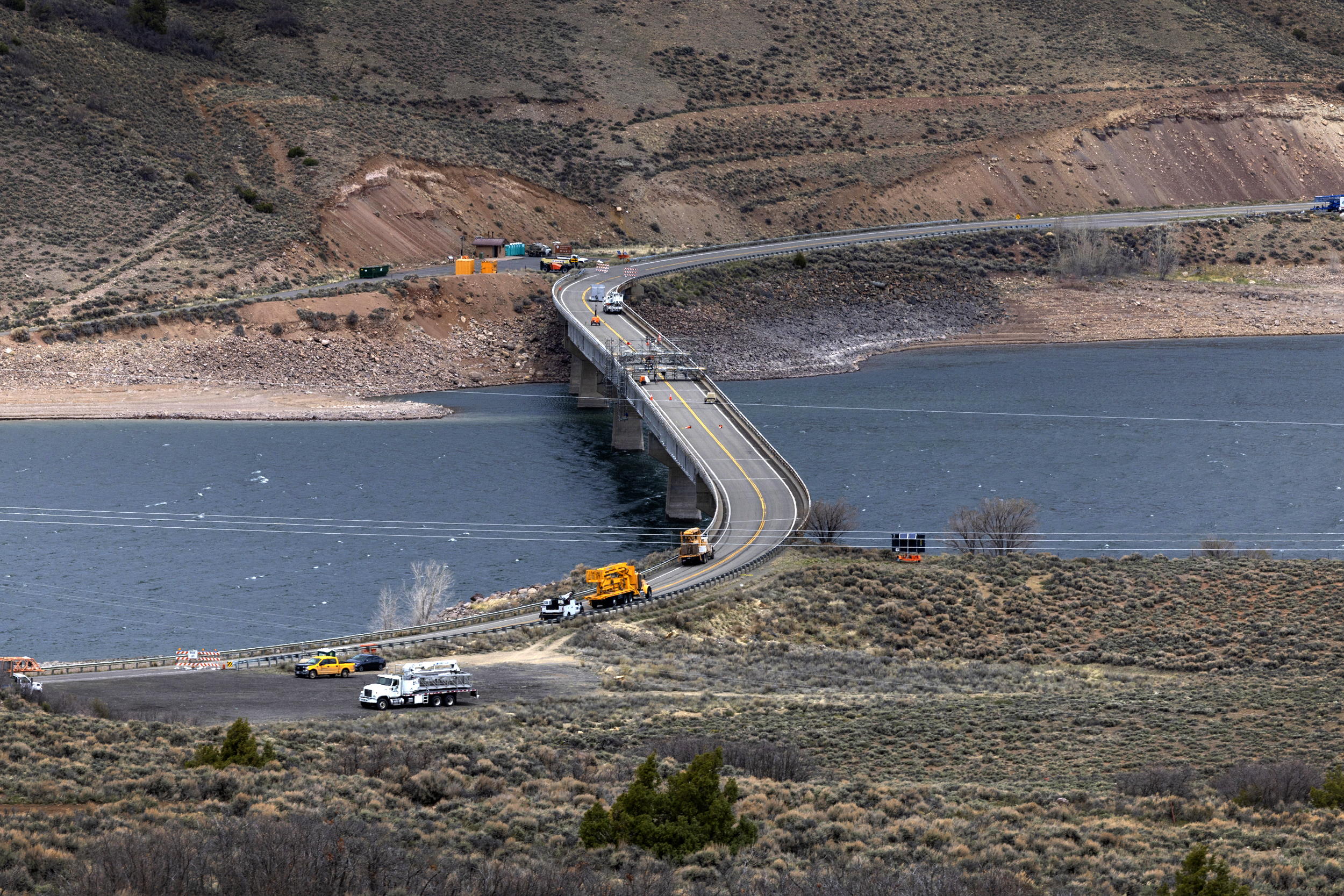 The U.S. 50 bridge under construction spans Blue Mesa Reservoir. Construction vehicles and equipment are on and near the bridge with machinery and traffic cones indicating ongoing work. Bushes and hills surround the area.