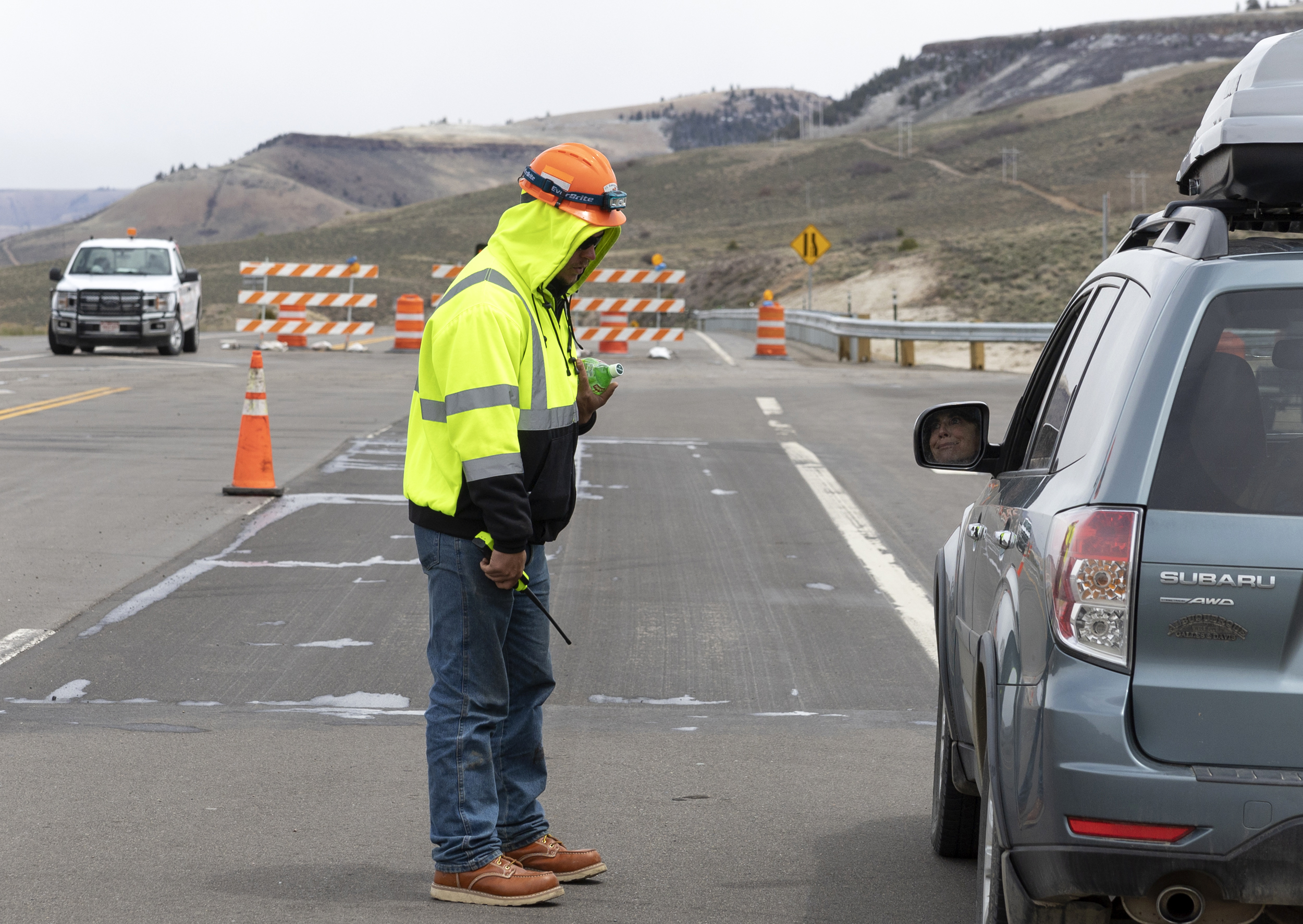 a man in neo green jacket and jeans with orange helmet stands next to a car on a highway