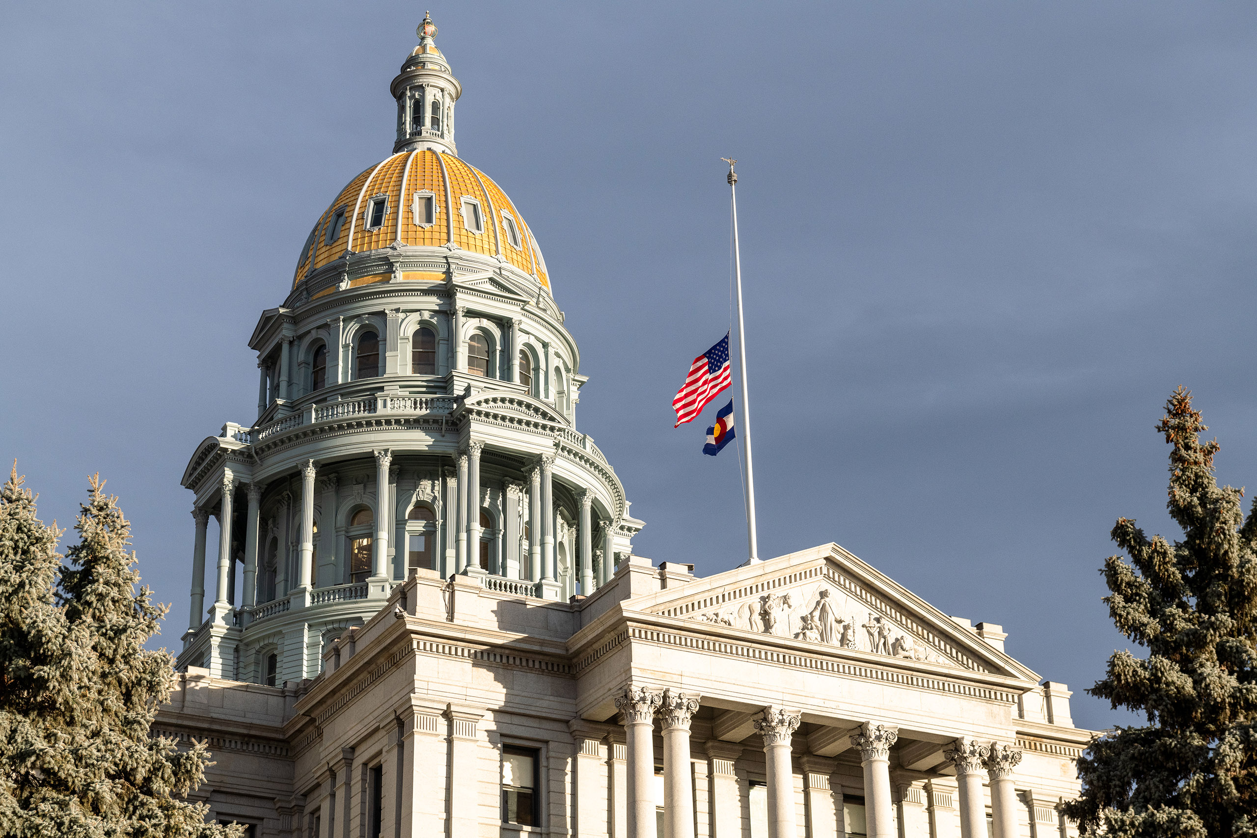 The Colorado State Capitol building with a gold dome and a flag at half-staff against a cloudy sky.