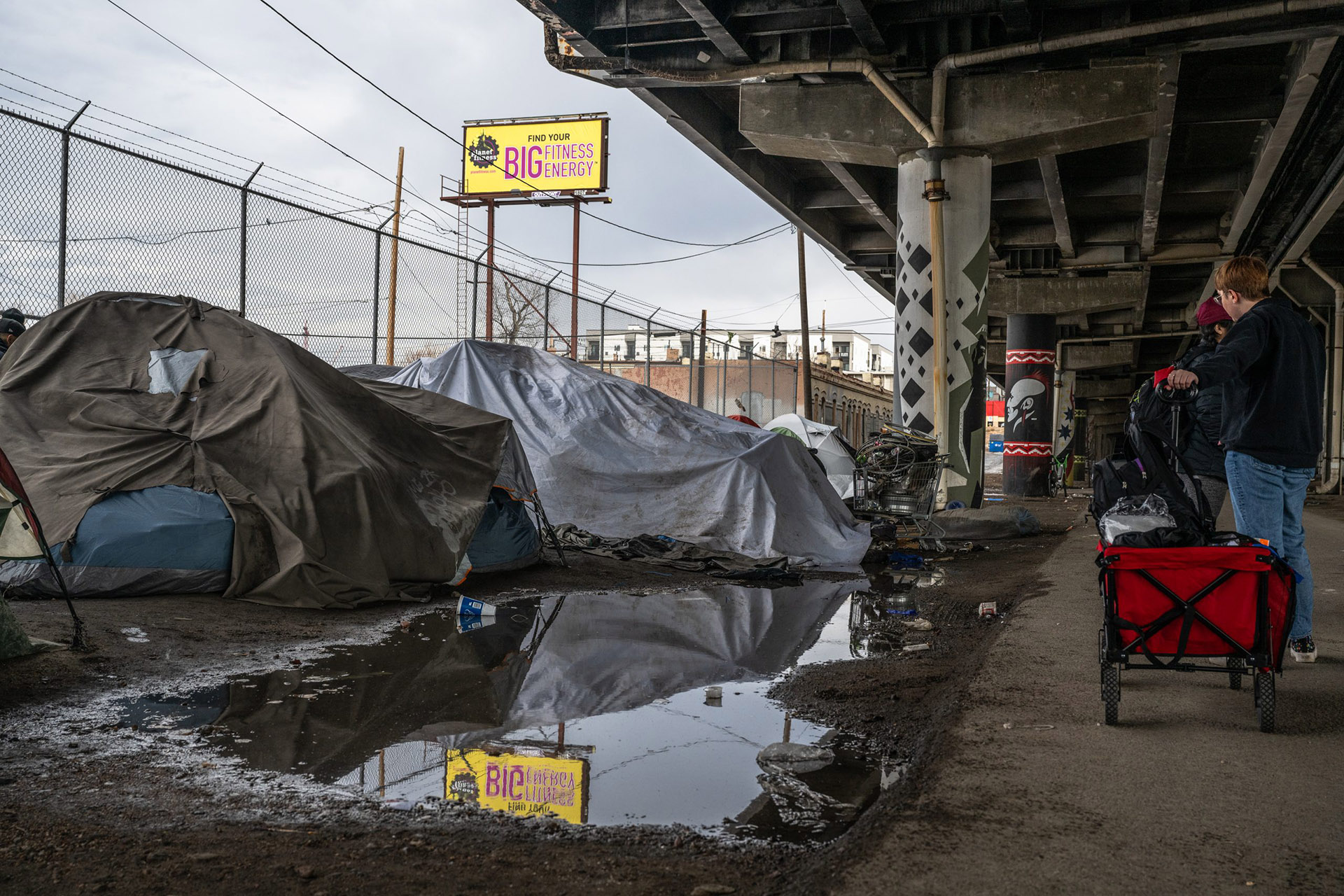 A person with a red cart stands near a row of makeshift tents under an overpass, highlighting the stark reality of homelessness. A large puddle on the ground nearby reflects a billboard for an energy drink in the background.