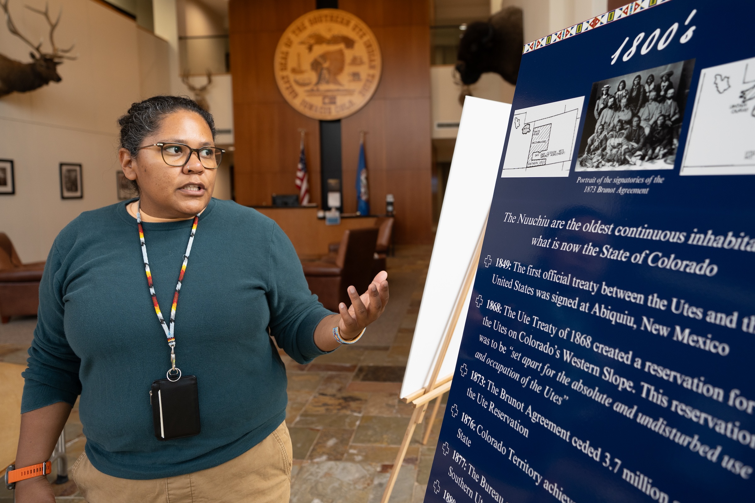 A person stands next to a display board on Ute history, gesturing while explaining. The board includes historical details about the Ute Tribe in Colorado.