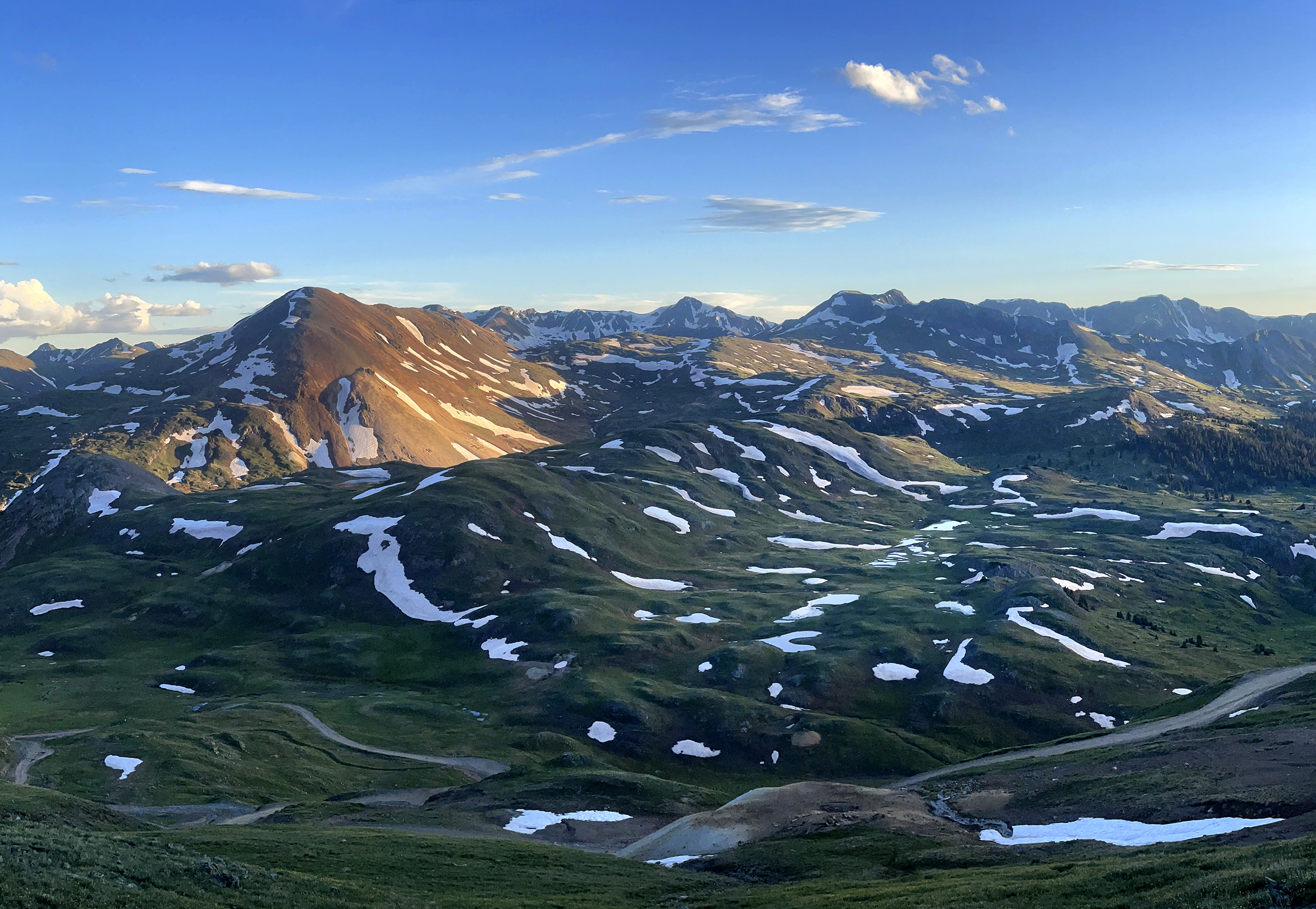 An aerial view of mountainous terrain with snowpacks in pockets across rolling green fields. A road cuts through.