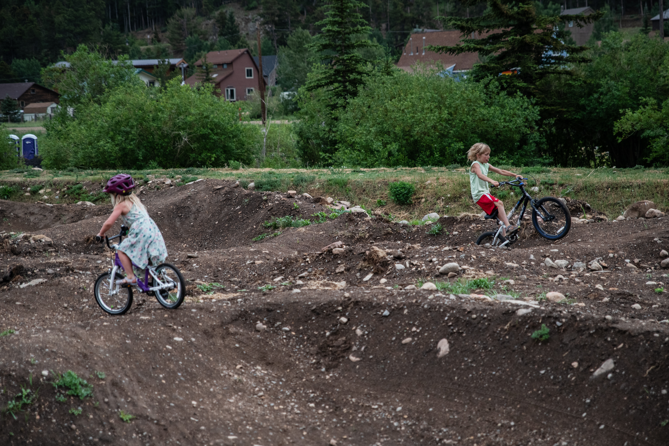 Two children ride bicycles on a dirt trail with small hills and curves. Trees, bushes, and houses are visible in the background.