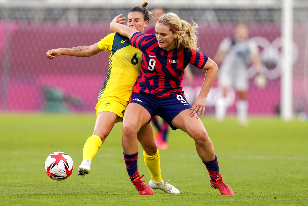 Two soccer players — Lindsey Horan and Chloe Logarzo — in action during a match. The player in yellow attempts to tackle the player in red and blue, who is holding the ball. The goalkeeper stands in the background near the goal.