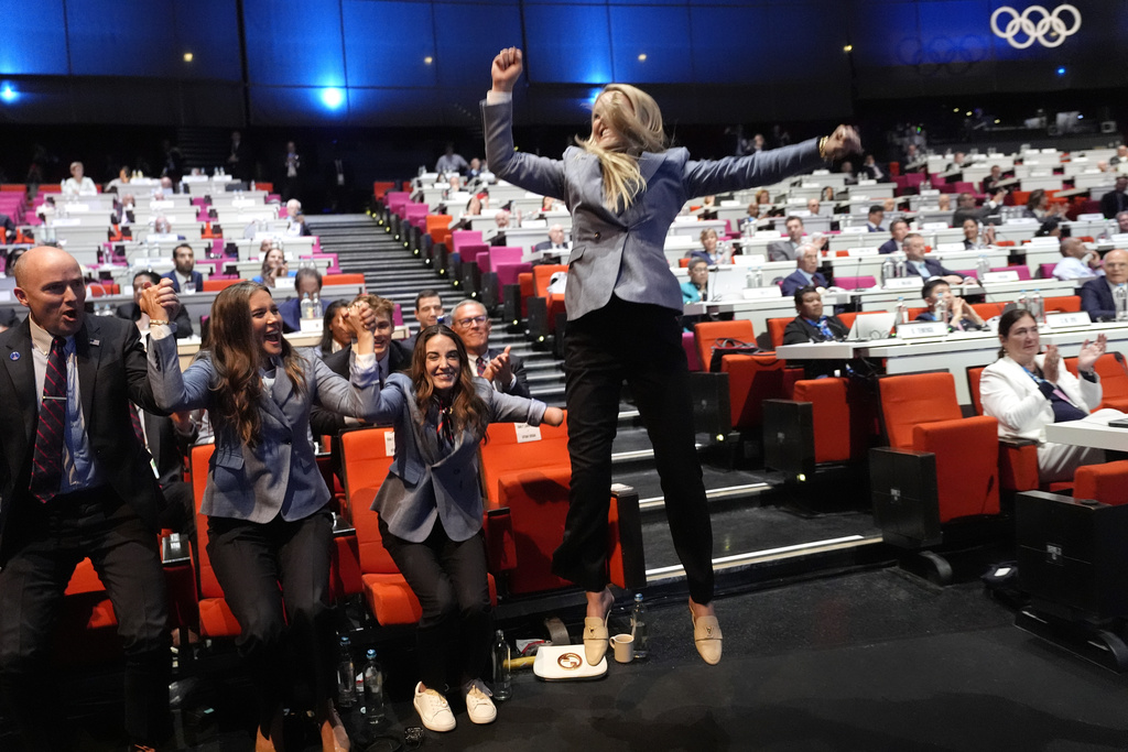 People in business attire cheer and celebrate at an event held in a large auditorium with an Olympic logo in the background.