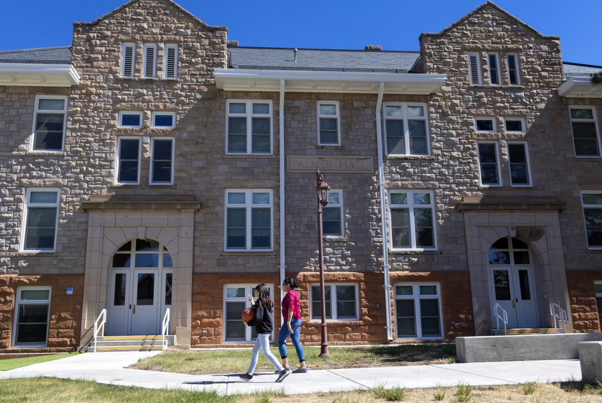 A pair of young people walk past large stonewalled building with windows on a sunny day.