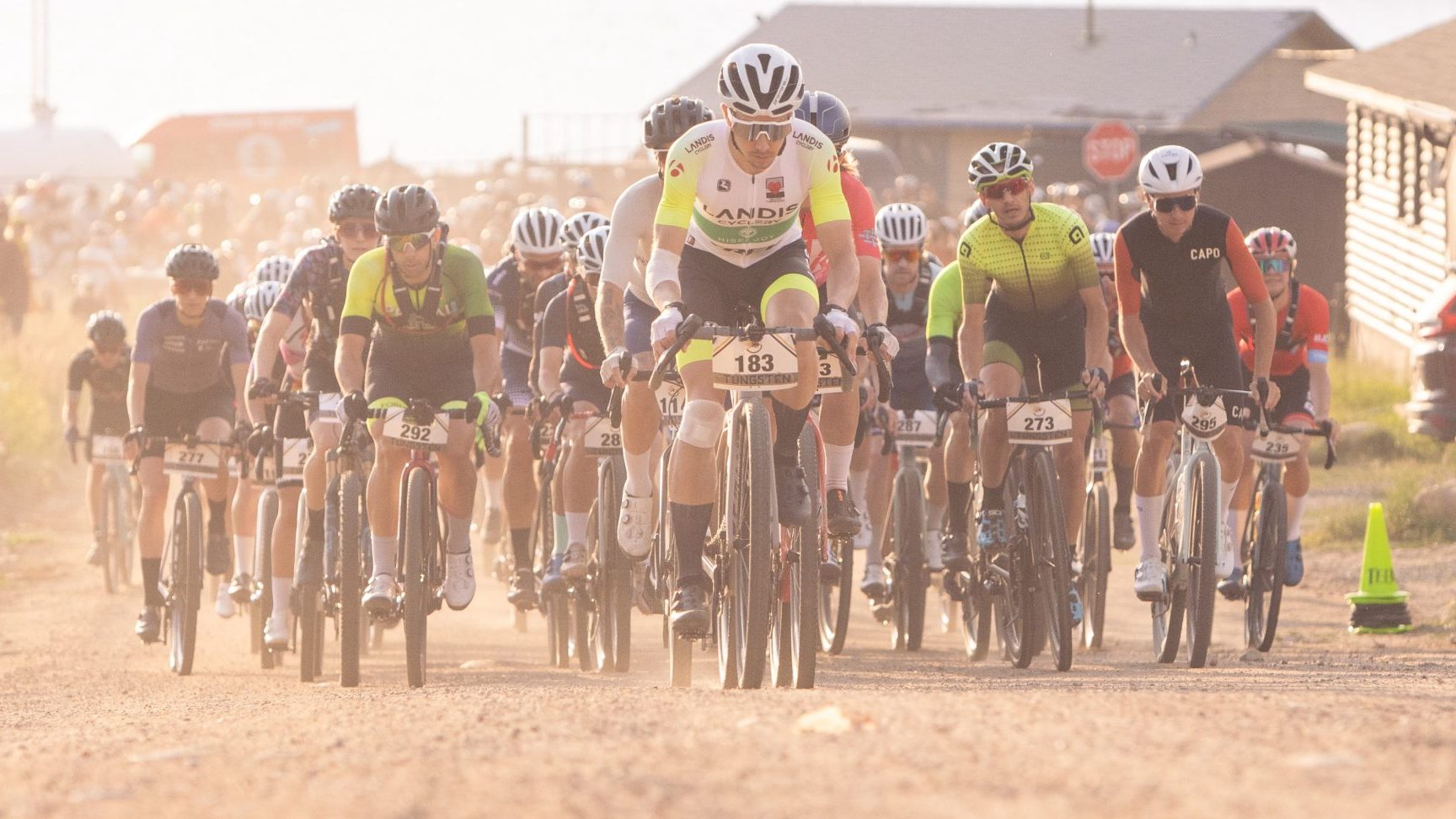 A group of cyclists ride on a dusty road during a race, with competitors wearing numbered jerseys and helmets. Buildings and parked cars are visible in the background.