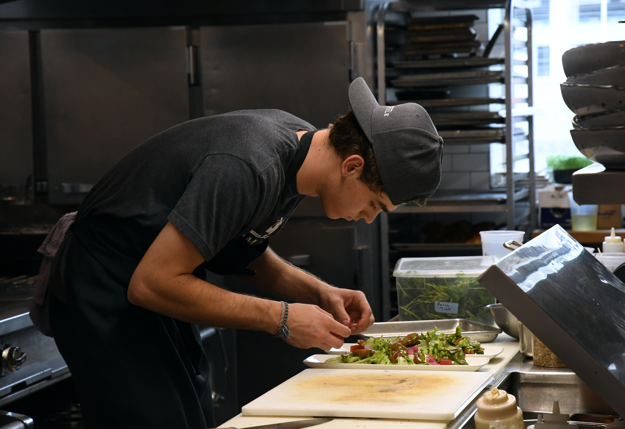 A person wearing a baseball cap and apron carefully prepares a salad in a commercial kitchen setting.