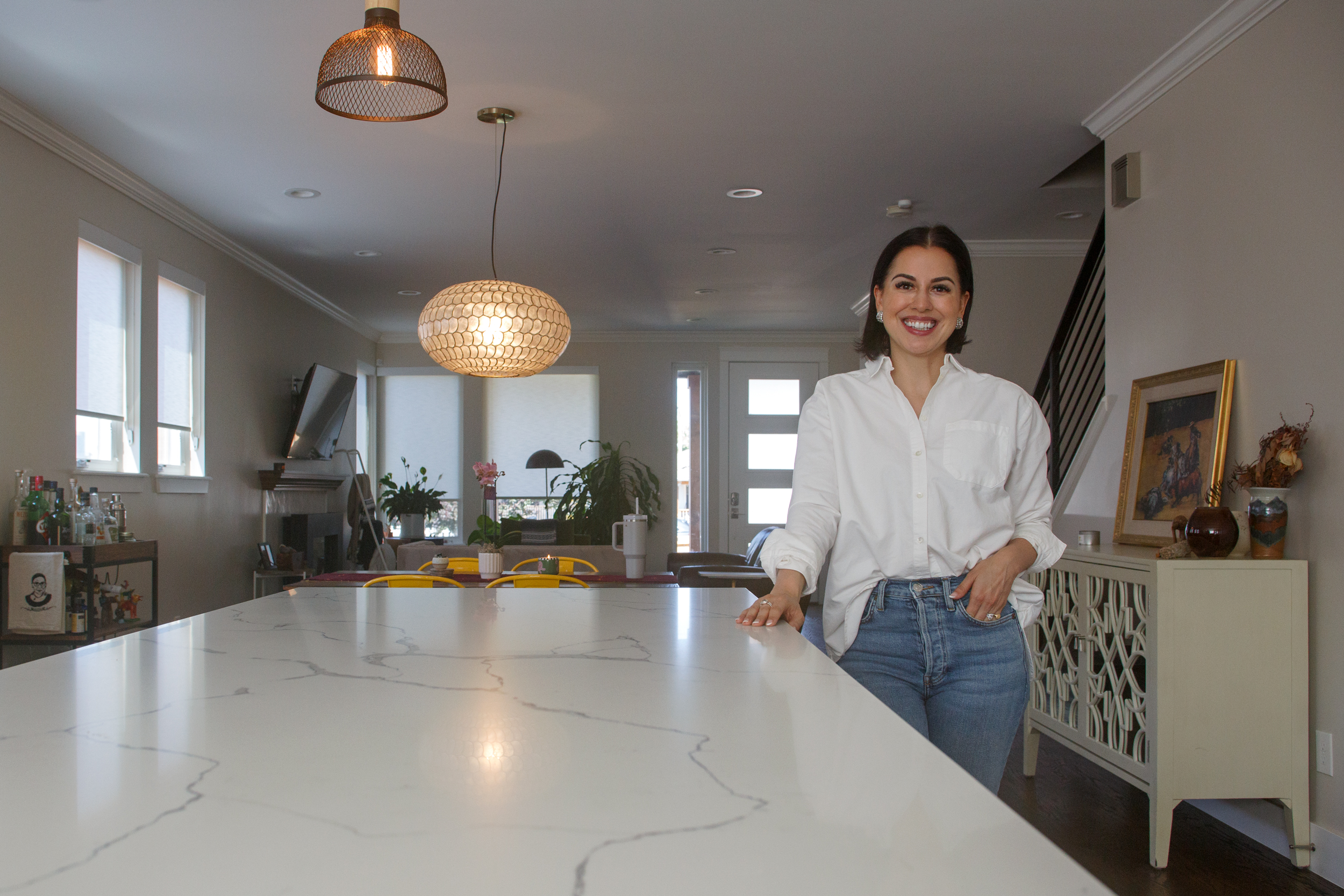 A woman in a white shirt and jeans stands by a marble kitchen island in a modern, well-lit kitchen-dining area.