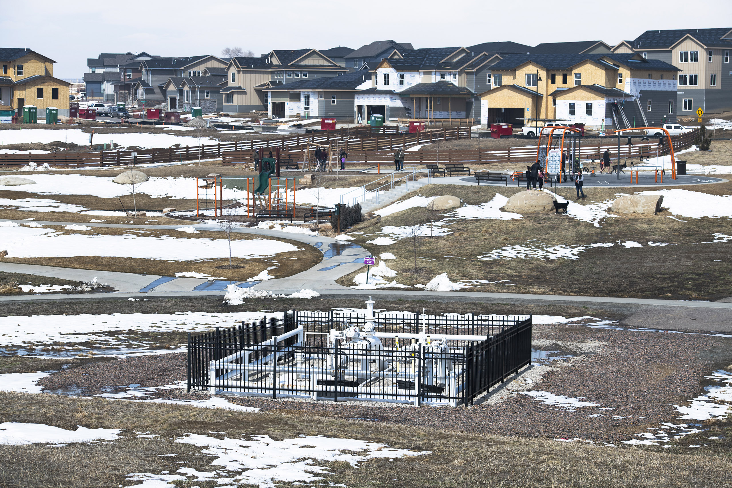 A suburban neighborhood showcases snow patches on the ground, scattered houses, a fenced-in utility area, and a playground with people in the distance. A cloudy sky overhead adds to the sense of air pollution.