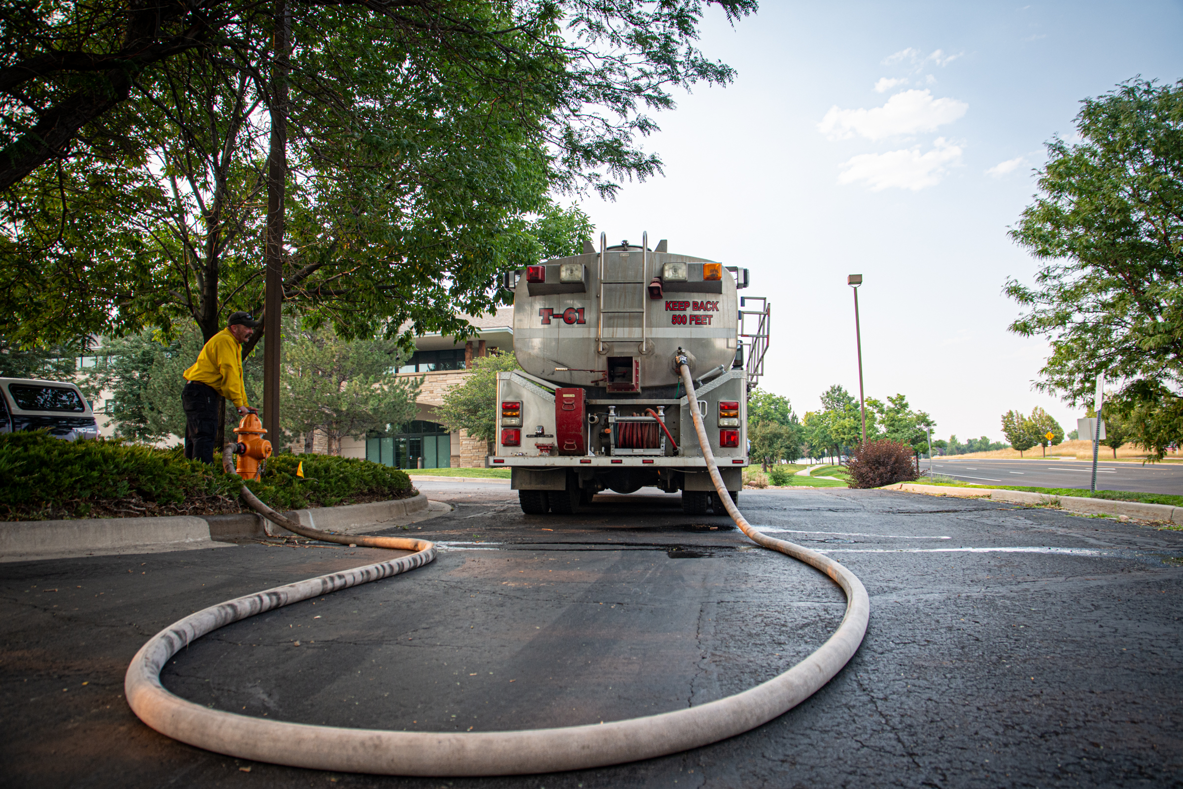 Worker filling a tanker truck with water from a fire hydrant on a tree-lined street.