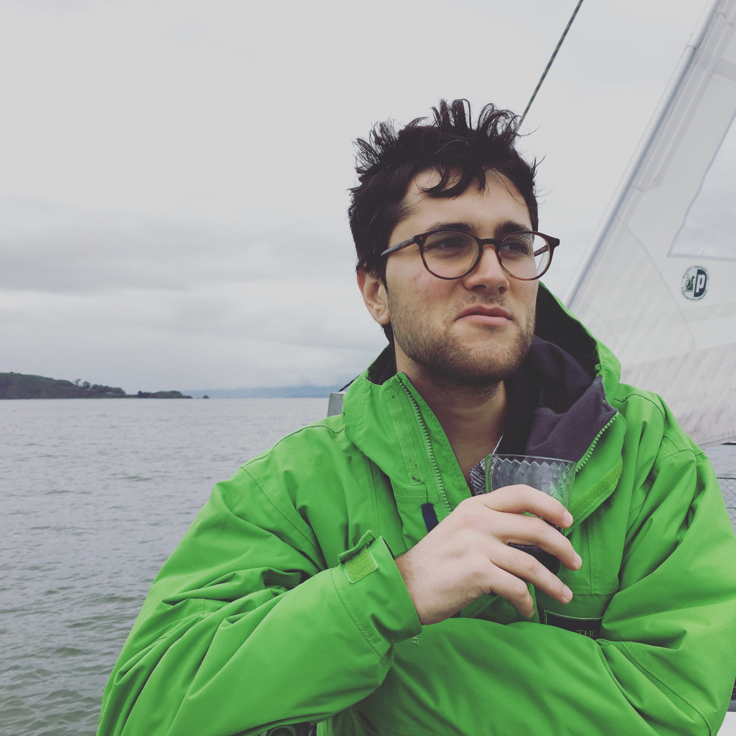 Man in green jacket and glasses sitting on a sailboat, looking pensively at the water, with overcast sky in background.