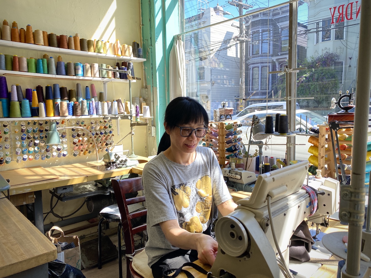 Shirley Xu sits behind the sewing machine with colorful thread spools in the back in Sunny Launderette.