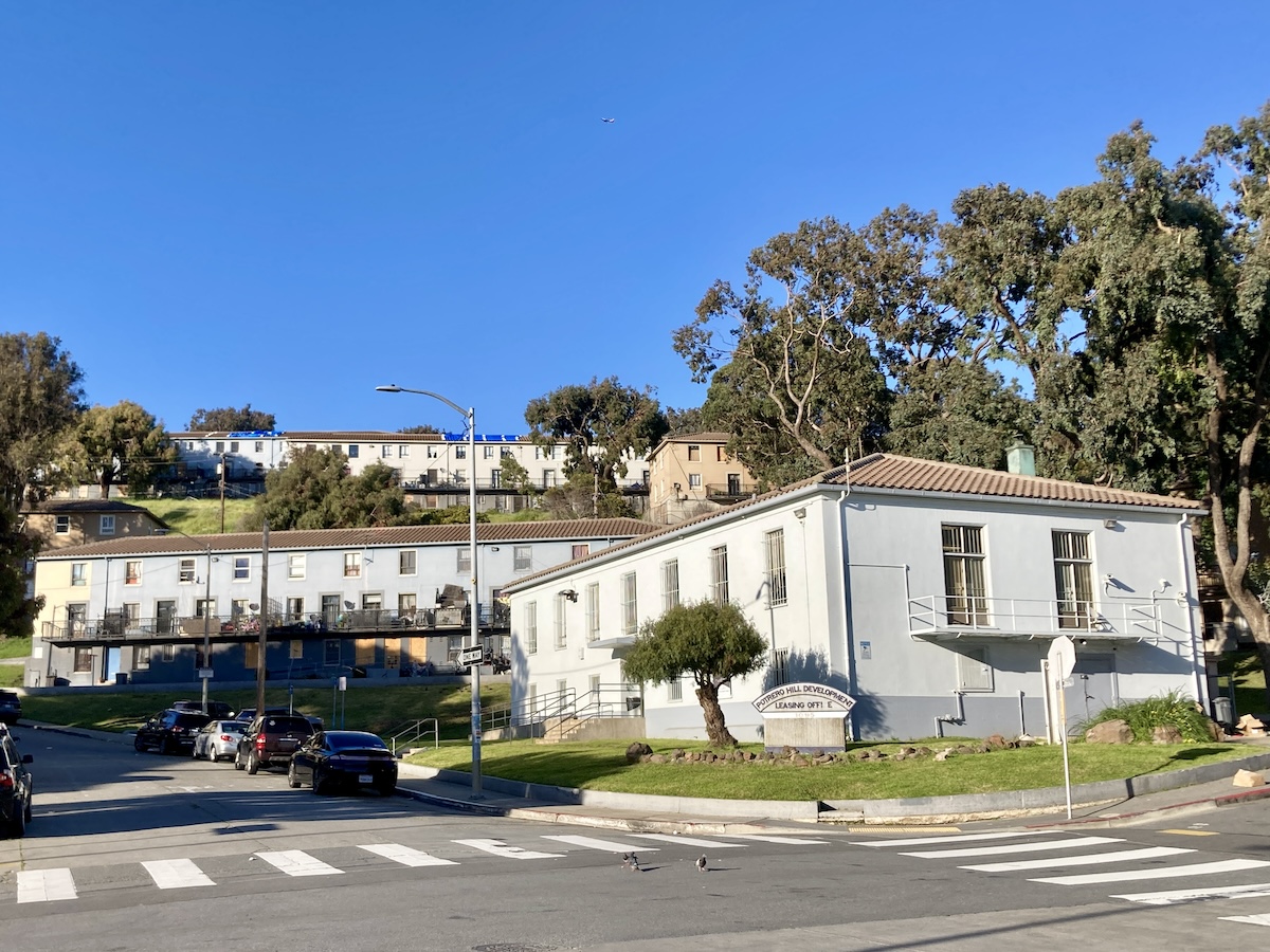 A sunny street view in Potrero Hill with a pedestrian crossing, cars, a gray building on the right with signage, and hillside homes in the background.