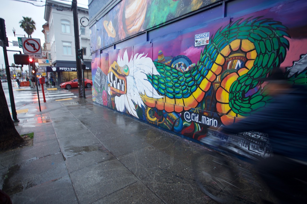 A colorful mural of a dragon on an urban street wall with a pedestrian passing by in motion blur.