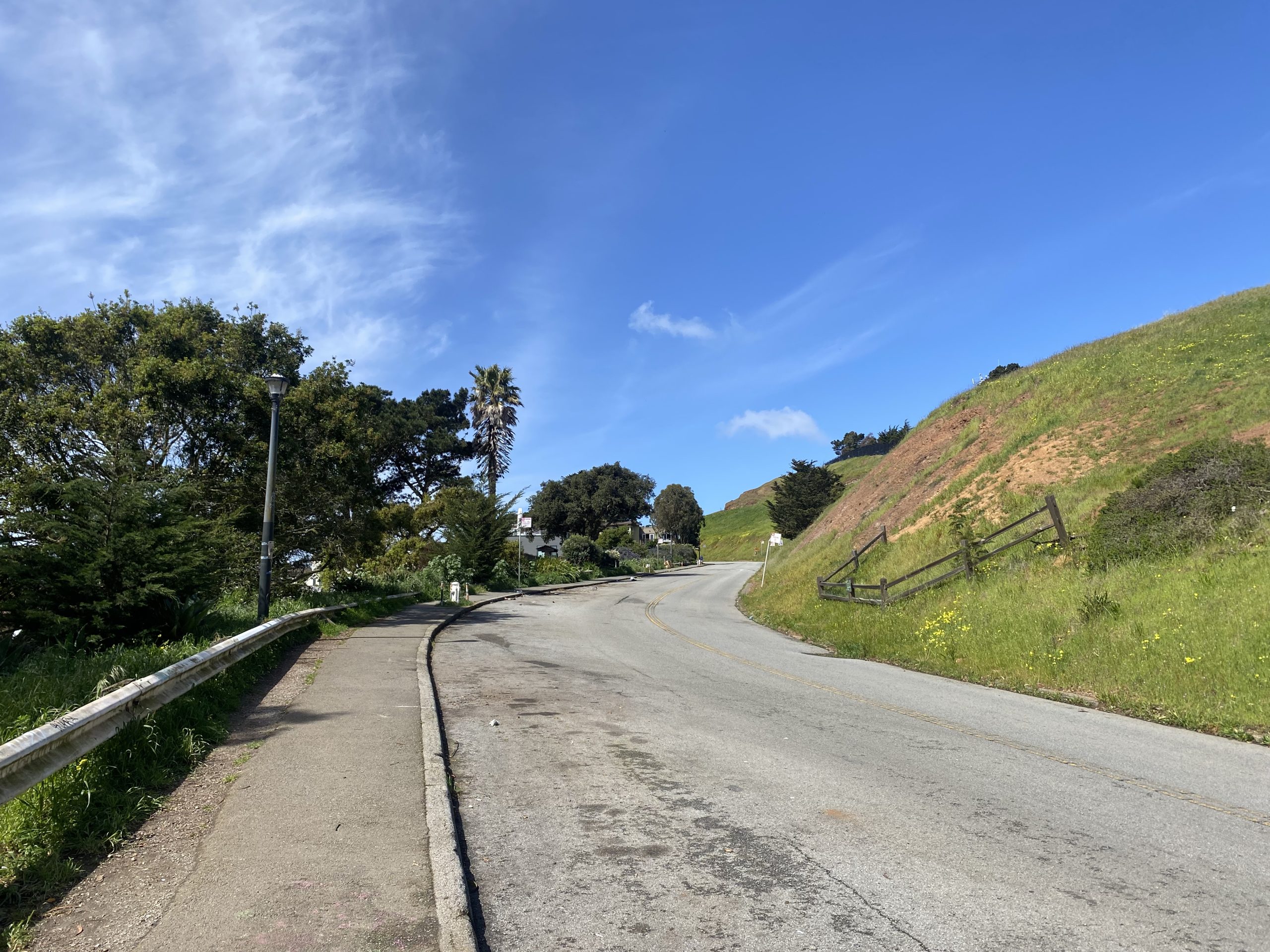 A winding road leading uphill with a clear blue sky, surrounded by greenery and a weathered guardrail on the left.