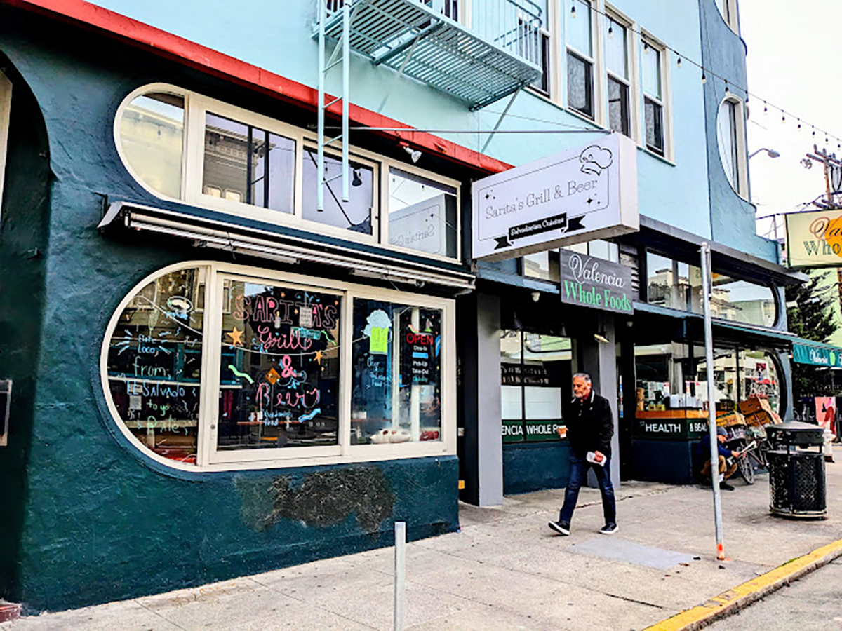 A pedestrian walks by a colorful storefront with a "grill & bar" sign and chalkboard menus visible through the window.