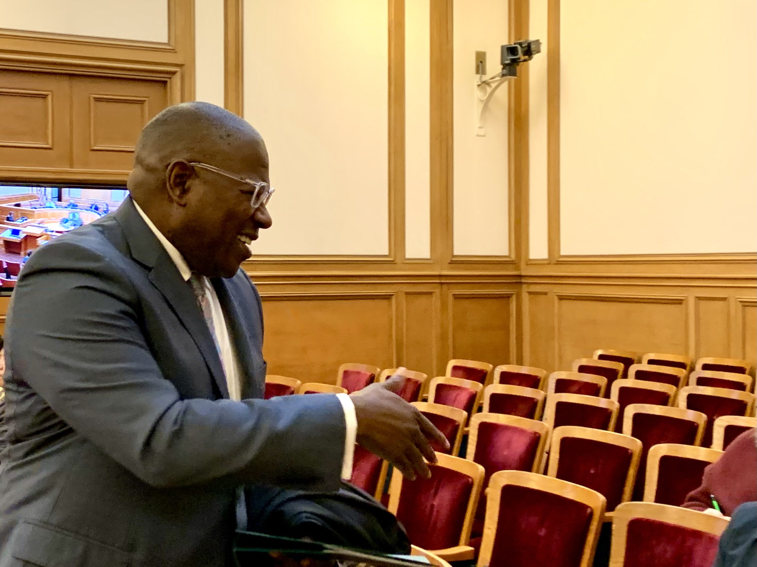 A man in a suit gesticulating during a conversation in a conference room with red chairs.