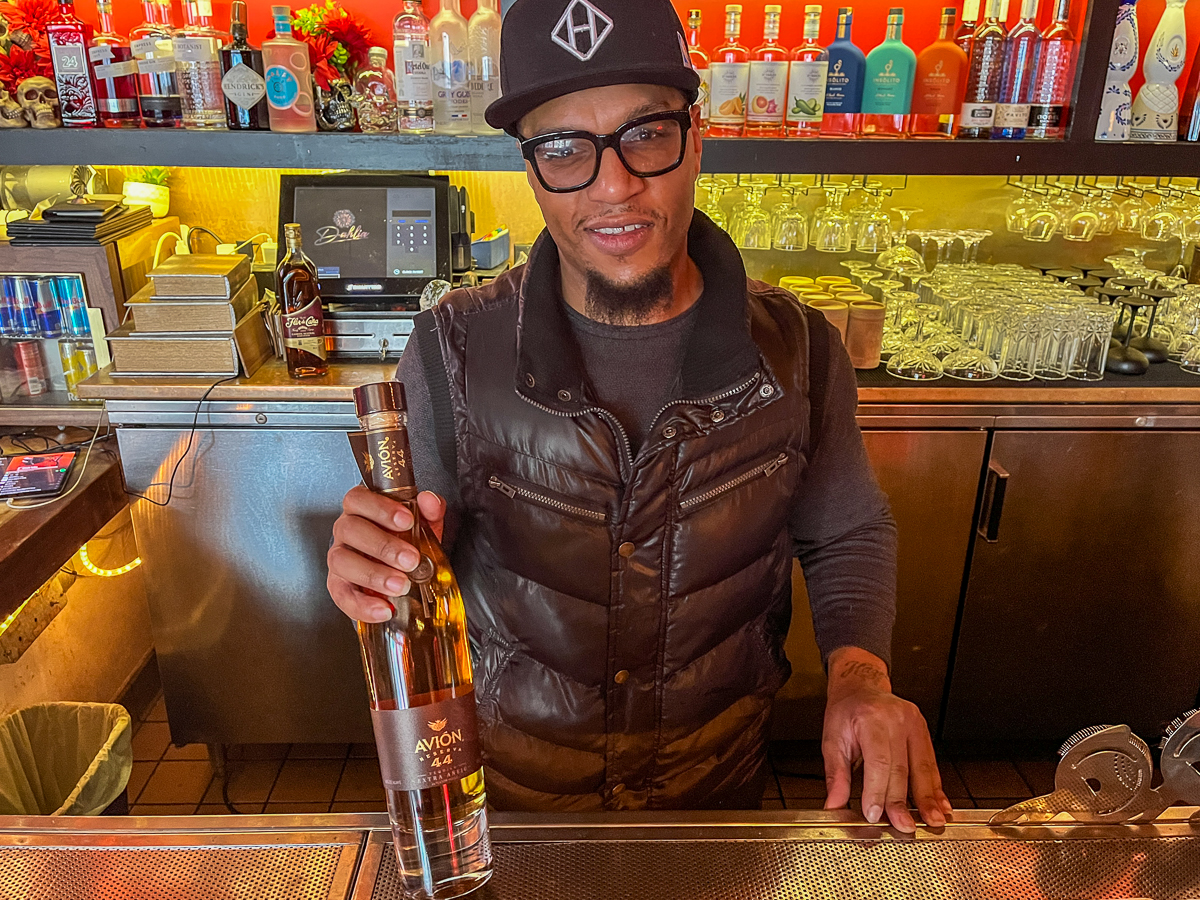 Bartender holding a bottle of avion tequila, smiling, standing behind the bar with shelves of various bottles in the background.