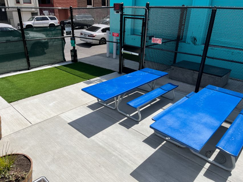 Outdoor seating area with two blue picnic tables on a concrete surface, adjacent to a fenced artificial grass area, with parked cars and buildings outside the fence.