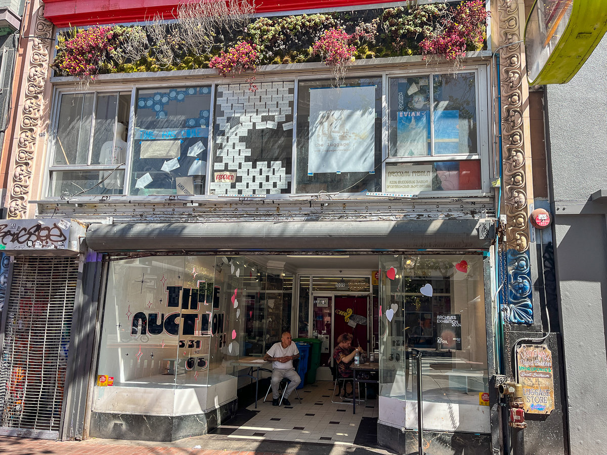 The image shows the storefront of a building with a "Time Auction" sign above the entrance. People are sitting inside and outside, and plants are hanging from the upper level.