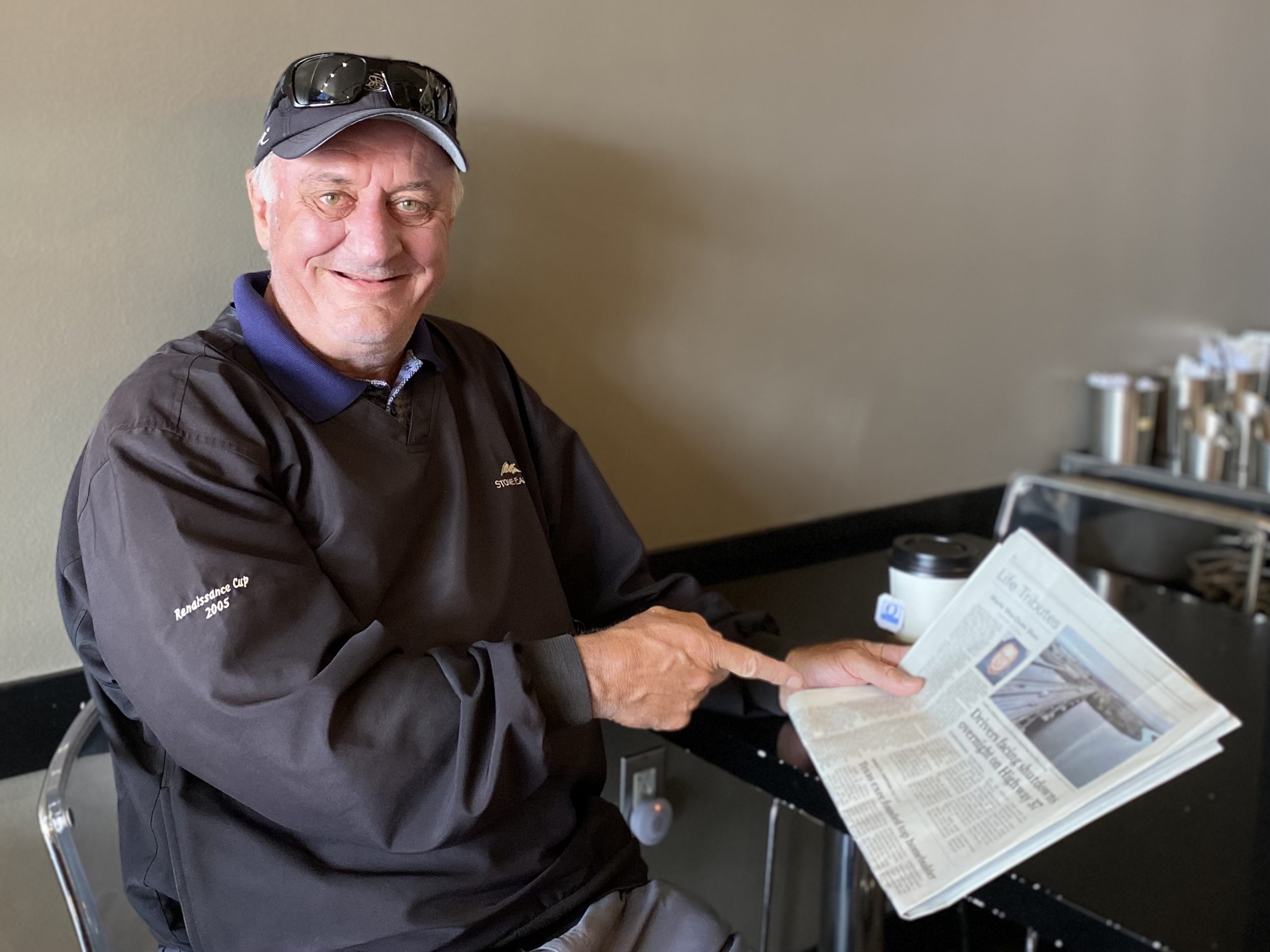 A man sits at a table, smiling while pointing at an article in a newspaper. He is wearing a dark jacket, a cap, and sunglasses on his head. A beverage container is visible on the table.