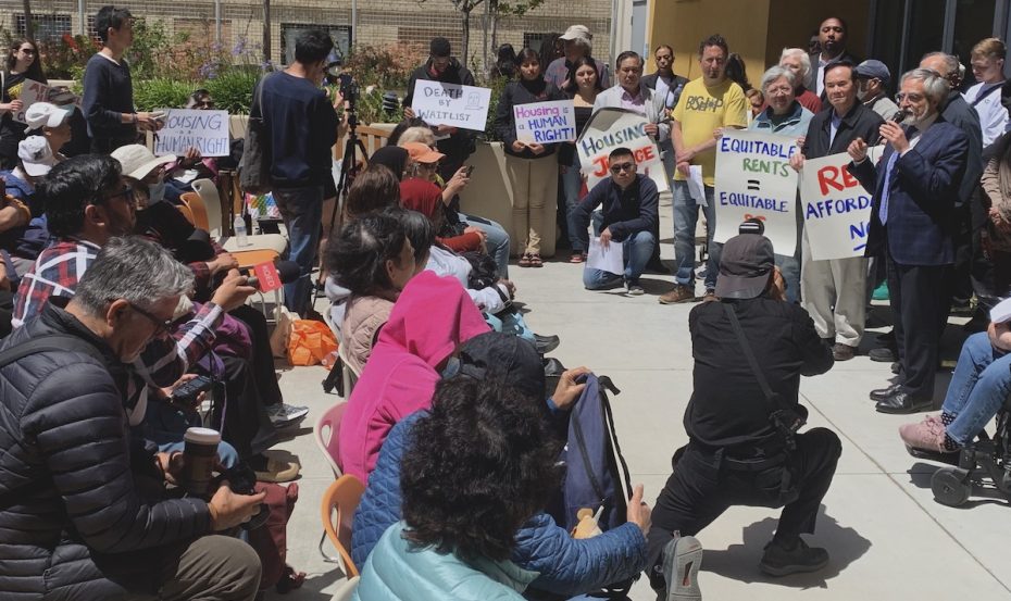 A crowd gathers at a rally, many holding signs with messages like "Housing is a Human Right" and "Equitable Rents." As the speaker addresses the group, attendees listen attentively. Peskin's name is frequently mentioned as an advocate for these pressing issues.