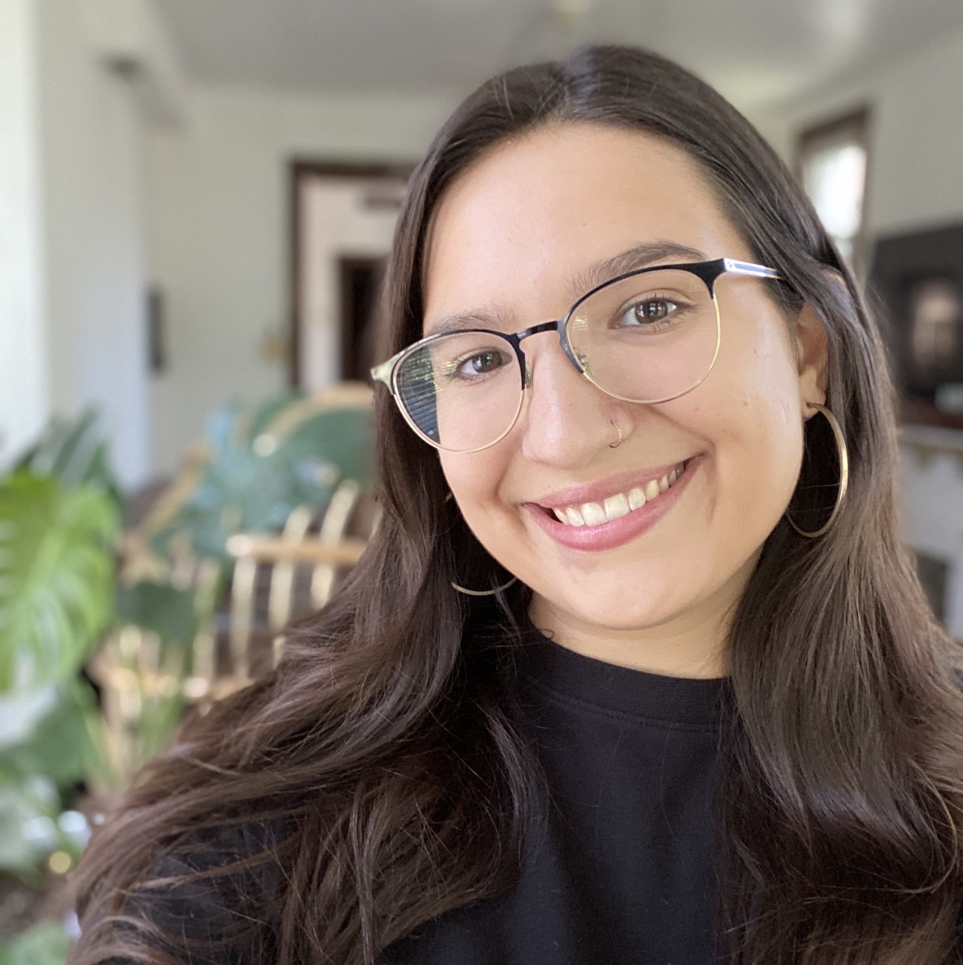 Young woman with glasses smiling at the camera in a room with green plants and furniture in the background.