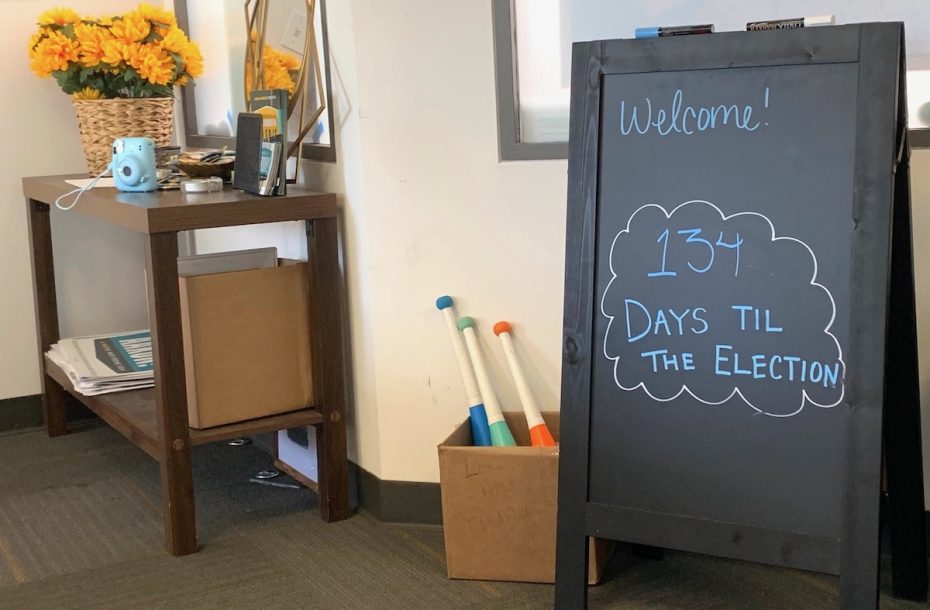 一间办公室的黑板上写着 "欢迎光临！距离选举还有 134 天。附近的一张桌子上摆放着一个花瓶，里面插着向日葵和各种物品，地上的一个盒子里放着交通锥。

