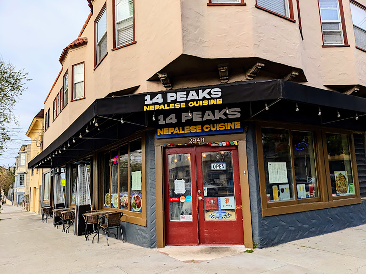 A corner restaurant named "14 PEAKS NEPALESE CUISINE" with a black awning. The red door has signs, and there are tables and chairs outside. The sign above reads "14 PEAKS NEPALESE CUISINE.
