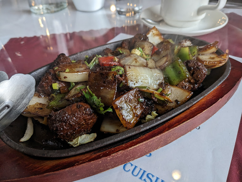 A sizzling dish of stir-fried beef, onions, bell peppers, and other vegetables served on a hot plate, accompanied by a stainless steel spoon.