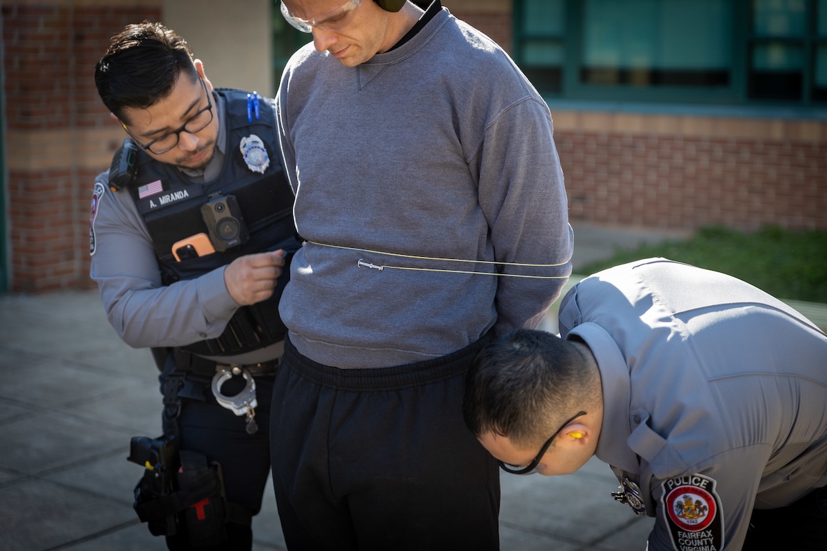 Two police officers are securing a man with zip ties. The man is wearing a gray sweatshirt. This interaction is taking place outdoors, in front of a brick building.