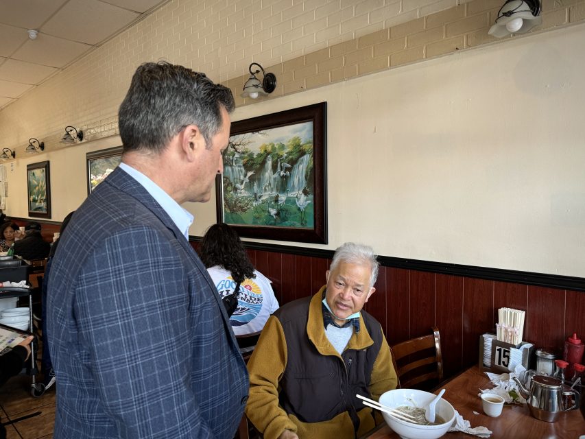 一位身着格子西裝的男子與坐在餐廳餐桌旁的一位年長男子交談，餐桌上的空湯碗、筷子和調味品清晰可見。