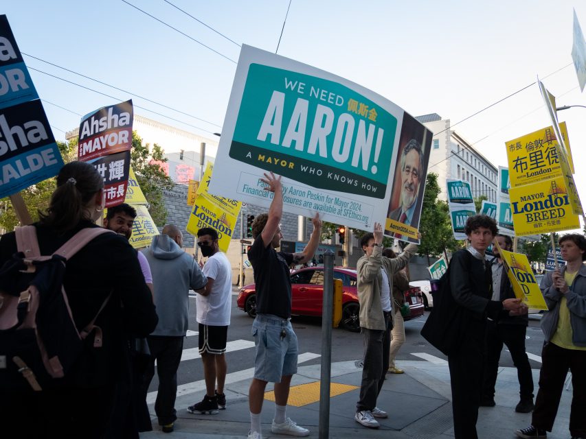 A group of people holding various political campaign signs, including "We Need Aaron! A Mayor Who Knows How" and "Ashade For Mayor," are gathered on a city street.