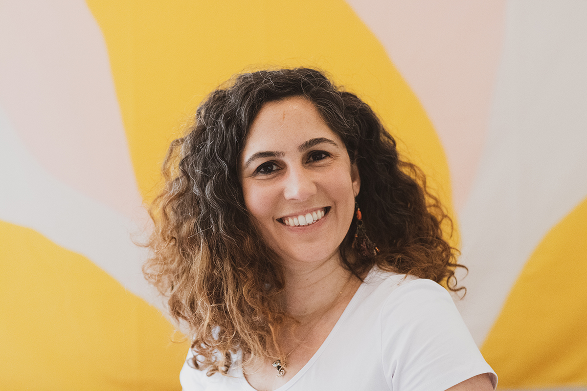 A person with curly hair, wearing a white shirt, smiles in front of a colorful yellow and white background.