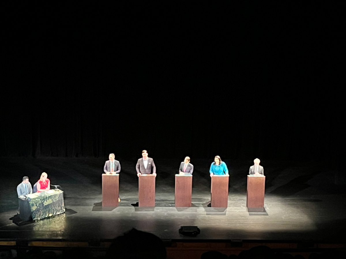 Five individuals stand at podiums on a stage, participating in a debate or discussion, while two people sit at a table to the side. The background is dark, providing contrast to the lit participants.