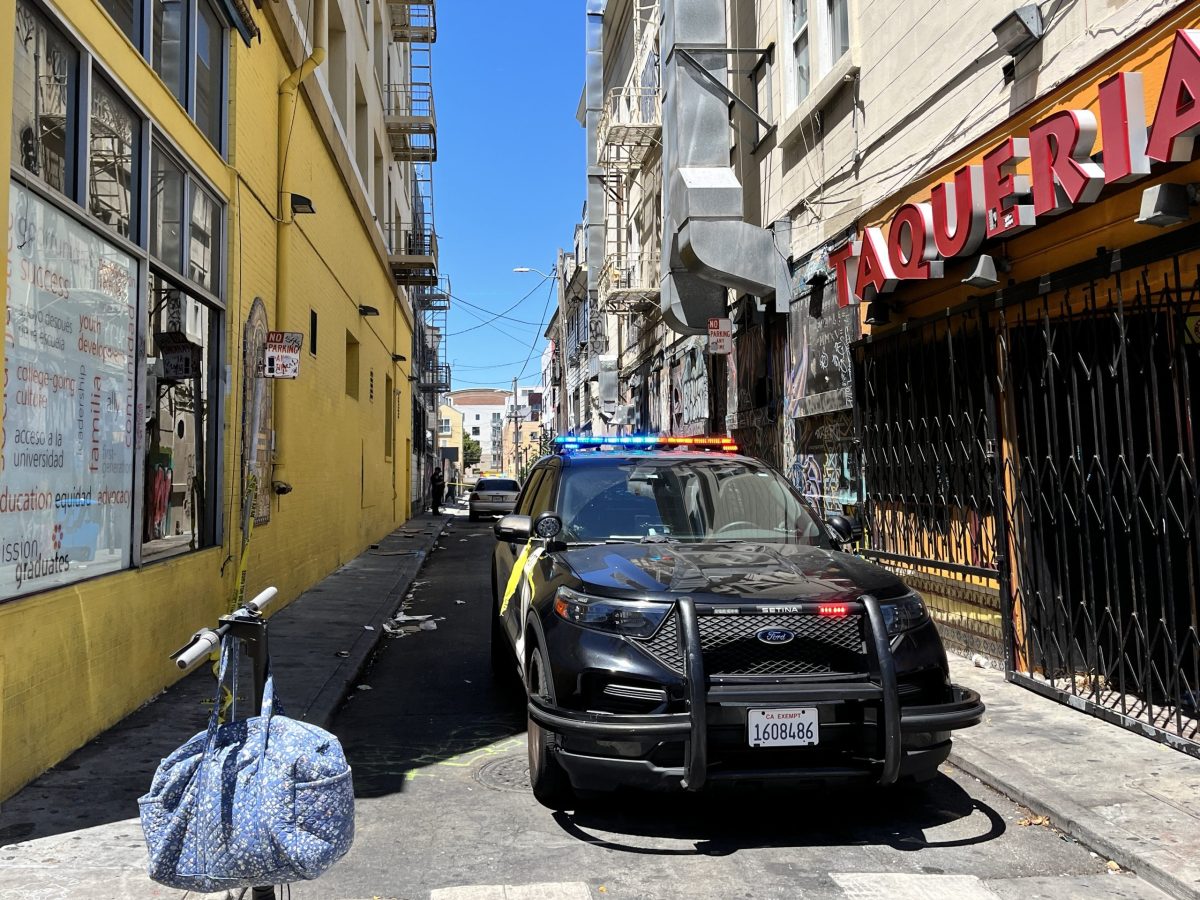 A police car with flashing lights is parked in a narrow alleyway next to a building with "TAQUERIA" signage. A bicycle with a blue bag is stationed nearby. Daytime scene.
