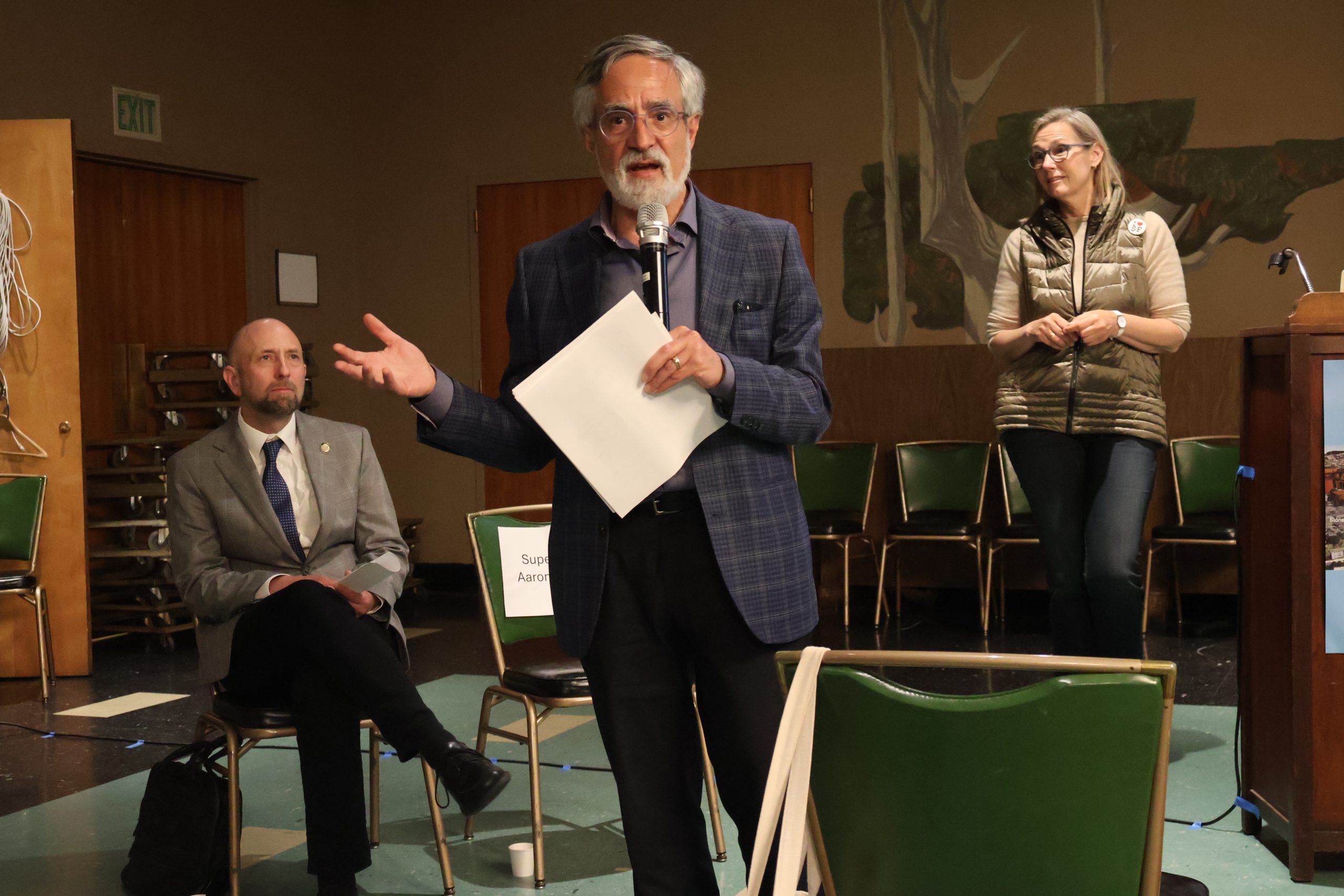 A man holding a microphone and papers speaks to an audience, with two individuals, one seated and one standing, in the background.