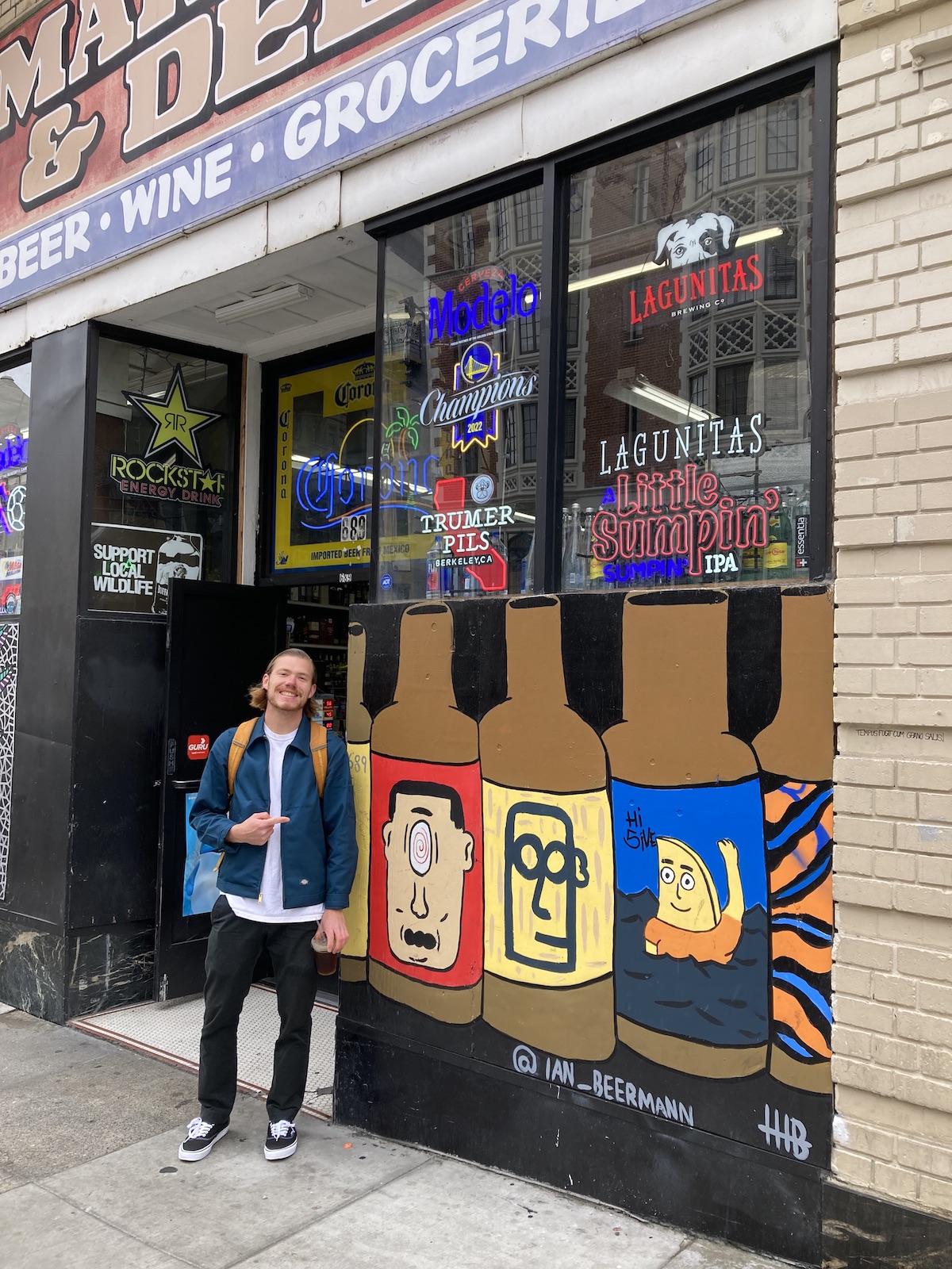 A man poses next to a mural of four cartoon bottles on the outside wall of a store with prominent beer and wine signs.