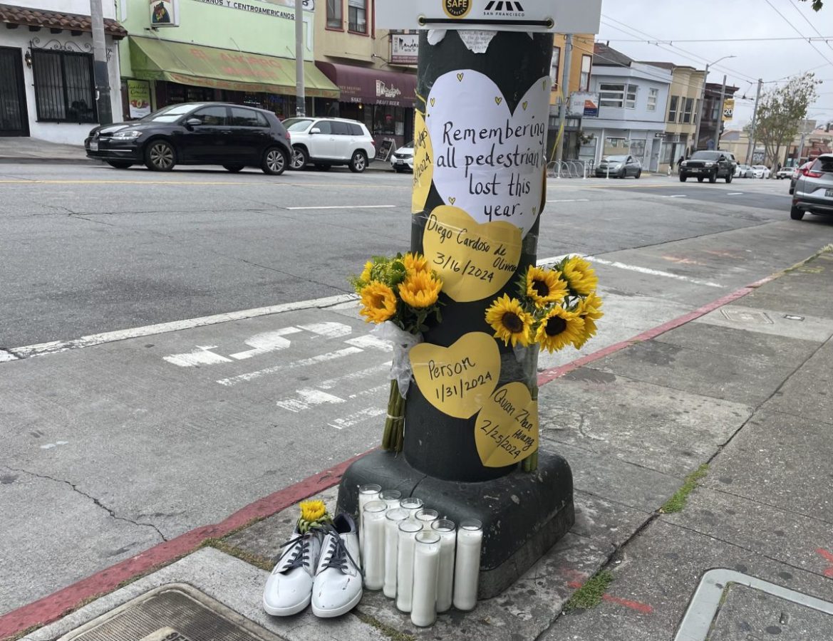 A street memorial with sunflowers, candles, and signs attached to a pole commemorates pedestrians lost in traffic incidents, featuring names and dates. A pair of white shoes and traffic on the street are visible.