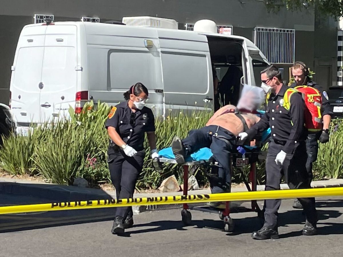 Emergency responders transport a shirtless person on a stretcher near a white van, while yellow police tape cordons off the area.