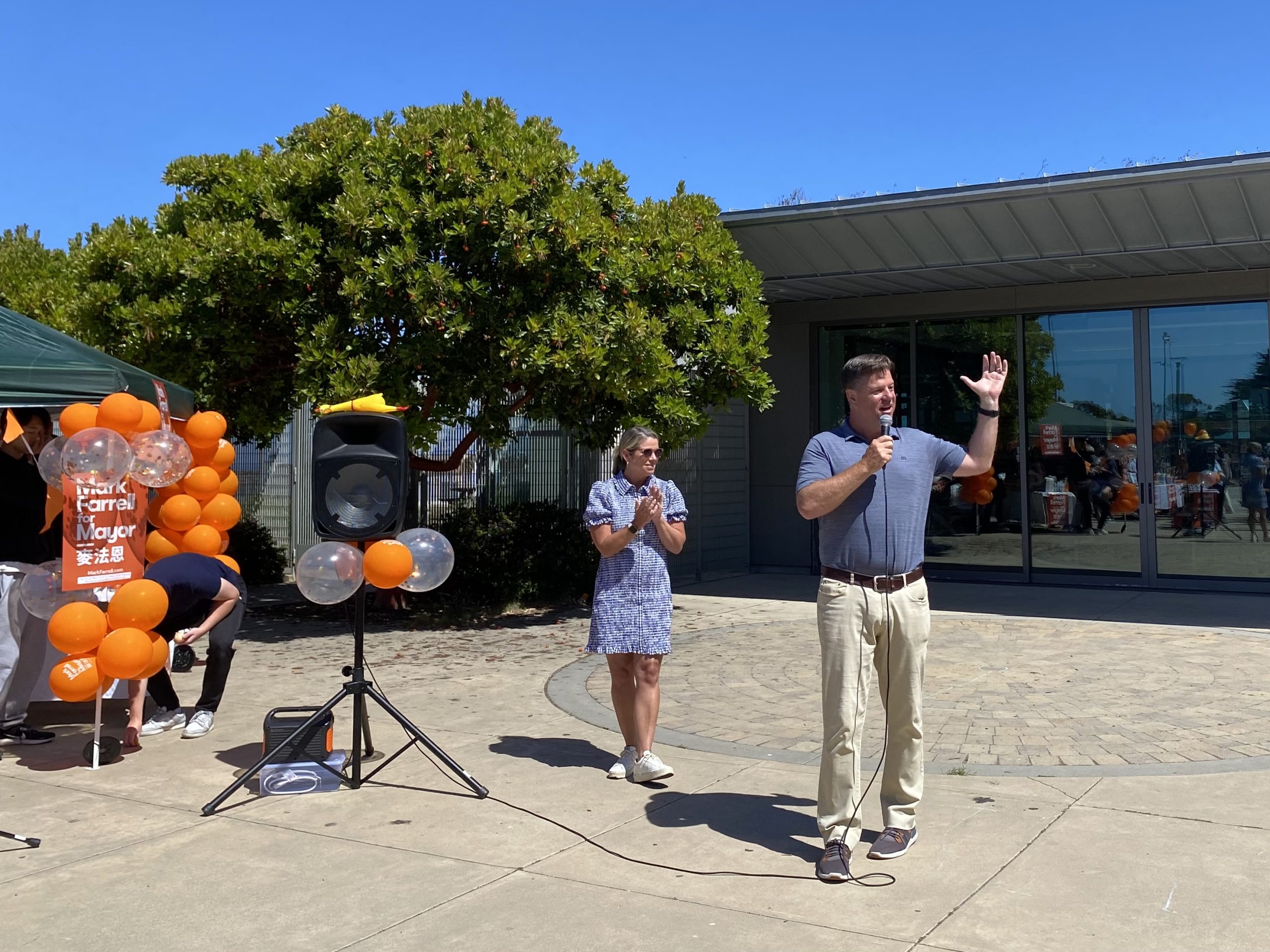 Mark Farrell speaks into a microphone, gesturing with one hand, while another person stands nearby clapping in front of a building. Orange and white balloons and a tree are in the background.