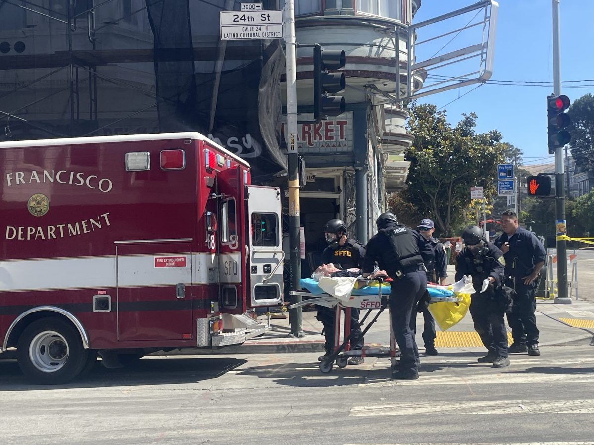 A group of police officers, including a SWAT team, and paramedics assist an individual on a gurney next to a San Francisco Department ambulance at an intersection in an urban setting.