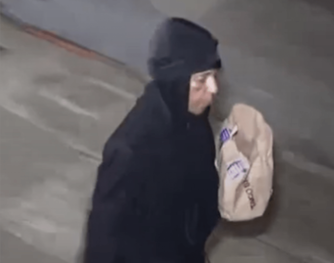 Person wearing dark clothing and a beanie walks on a concrete surface while carrying a beige paper bag with text on it. They are a person of interest in hate crimes against a local dog walker.