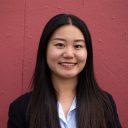 A person with long dark hair wearing a white shirt and black blazer stands in front of a textured red wall, smiling.